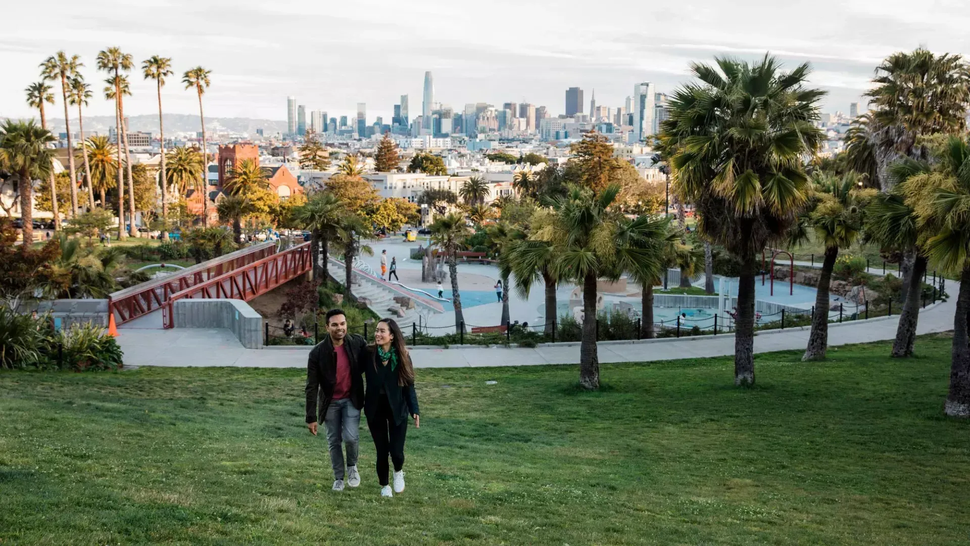 Una coppia cammina verso la telecamera, con Dolores Park e lo skyline di San Francisco alle spalle.