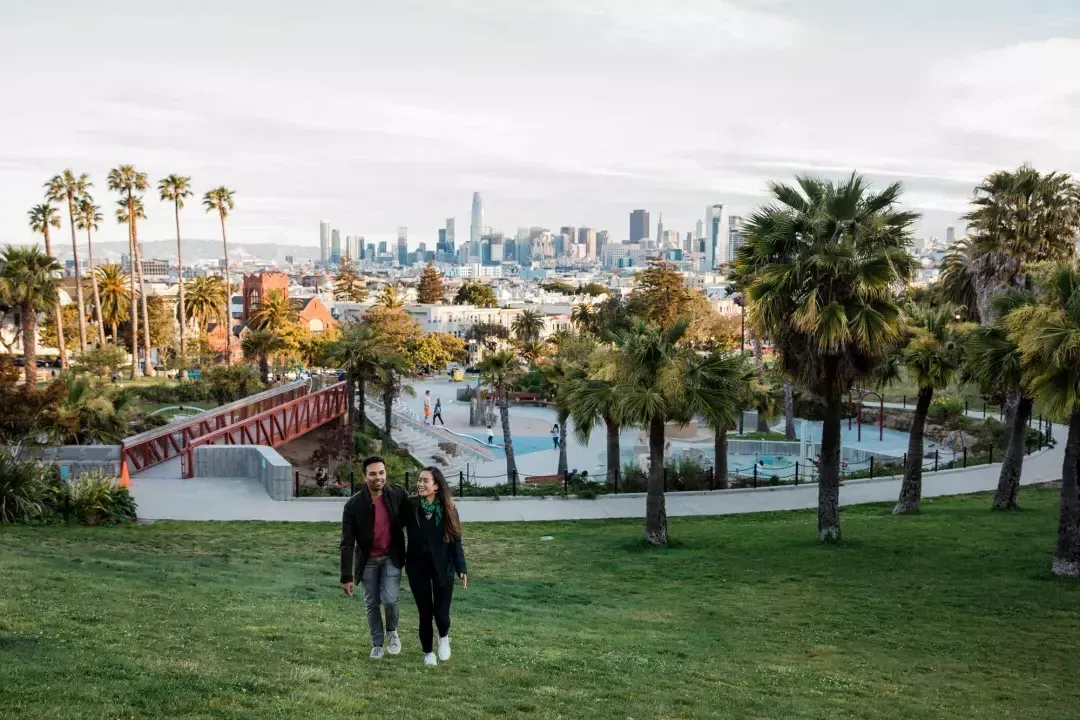 한 커플이 Dolores Park 과 샌프란시스코 스카이라인을 뒤로 한 채 카메라를 향해 걸어가고 있습니다.